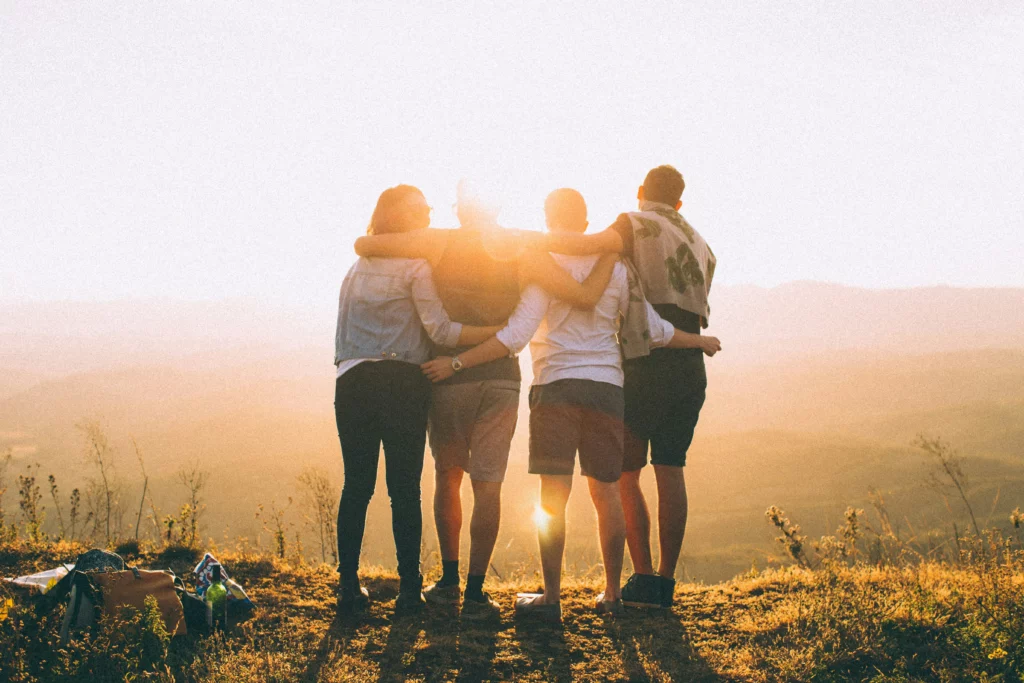 Four people holding on to each other, side-by-side while facing the sun and facing away from the camera.