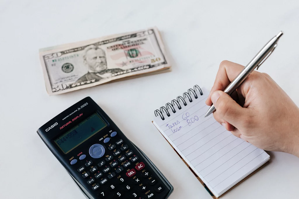 A stack of currency bills, the top one being a fifty-dollar bill lies on a white surface. A calculator is shown with numbers on its display screen. A hand holds a pen as it jots down expenses on a small spiral notebook. Hand-written text on notebook says taxes 60, loan 800.