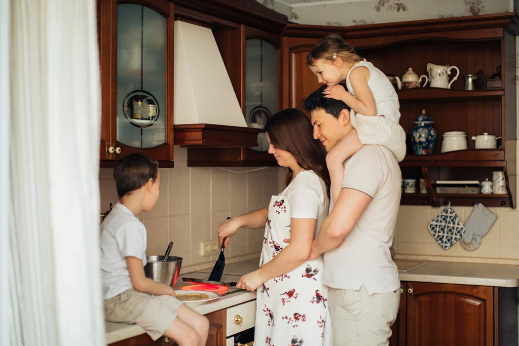 Familial unit of two adults and two children in a kitchen; the woman wearing an apron is cooking dinner to save money. The adult man holds her and is looking over her shoulder and at the cooking pan while having a child sit on his shoulders. A slightly older child sits on a kitchen surface and looks at the stove.