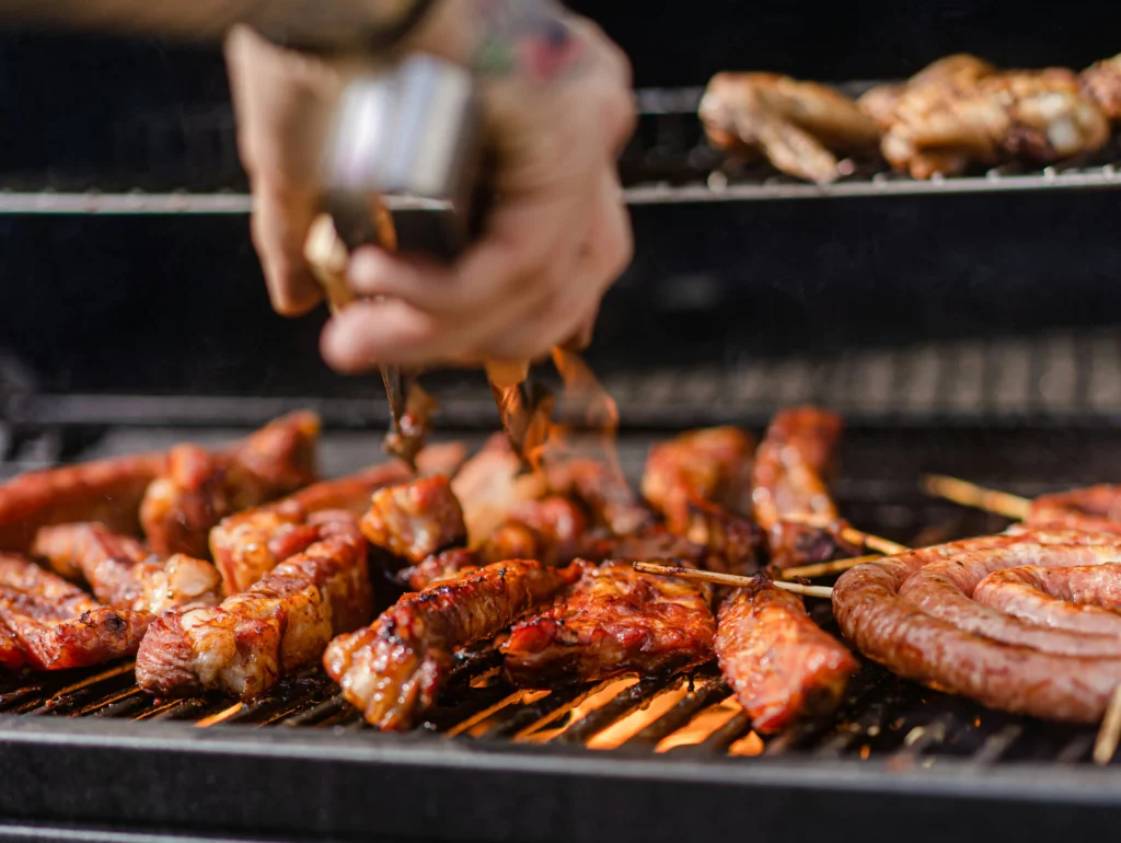 Person grilling barbeque for a wedding. A person is holding tongs and grilling up some meat. There’s chicken, sausage, and beef tips.