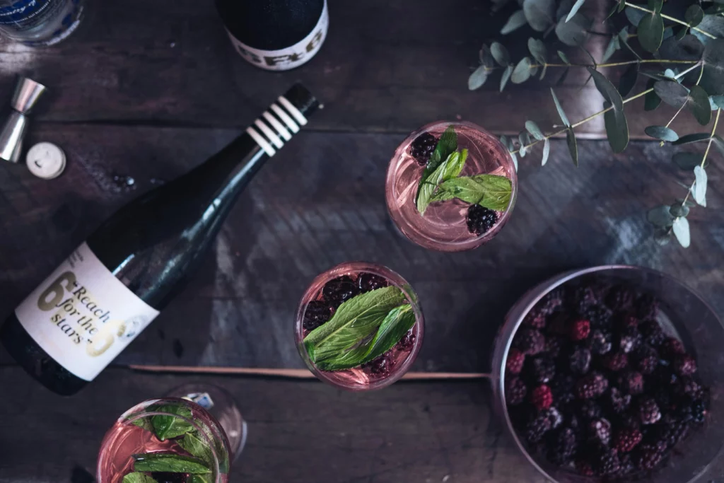 Signature cocktail at a wedding. Three glasses are on the table with a cocktail that includes mint and blackberries. The the left of the drinks is a bottle of sparkling wine. To the right of the drinks is a bucket of blackberries.