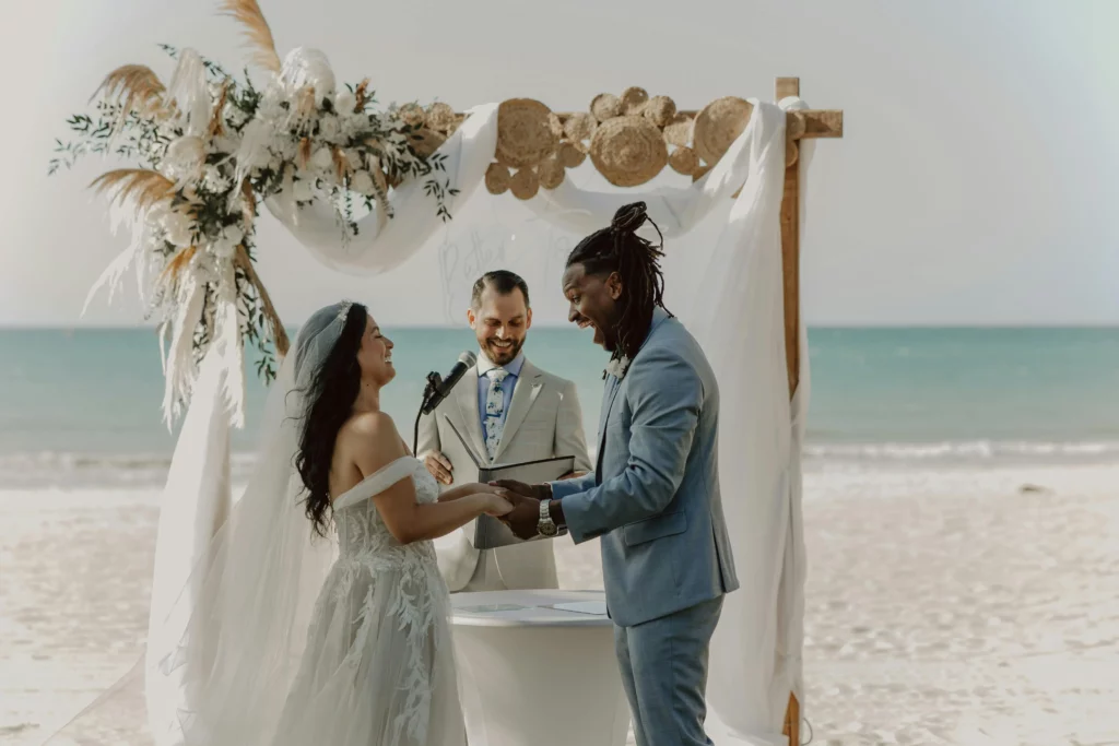 Couple laughing in front of wedding officiant. Couple getting married in a beach wedding They’re standing in front of their officiant who is laughing alongside them at the wedding altar.