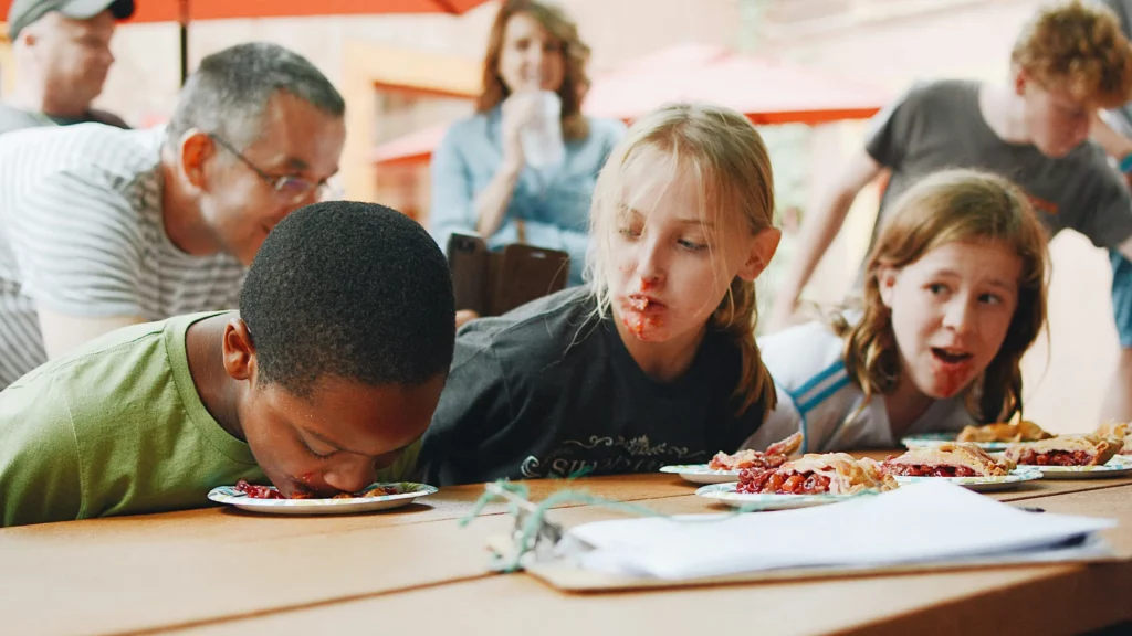Kids participating in a pie-eating contest on the 4th of July. Young child eats pie off of a plate without using his hands while two other children look up from their pies and at the first kid.