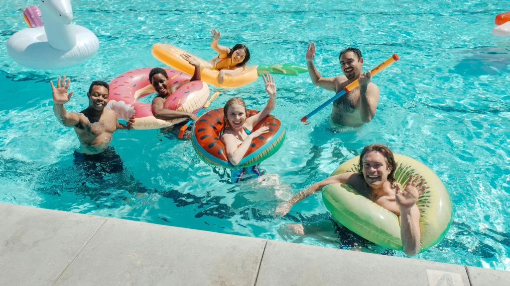 Six people having fun in a pool with several floatable tubes, smiling and waving at the camera.