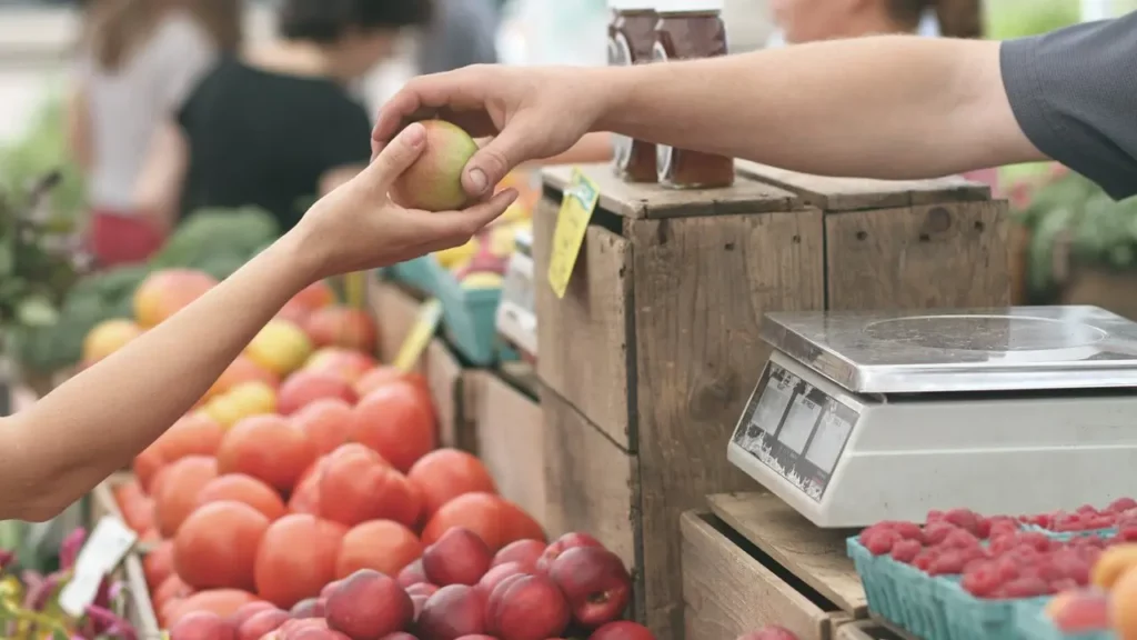 One hand giving away a fruit while another hand grabs to receive it. The fruit stand is in the middle of a farmers' market for local and small business.