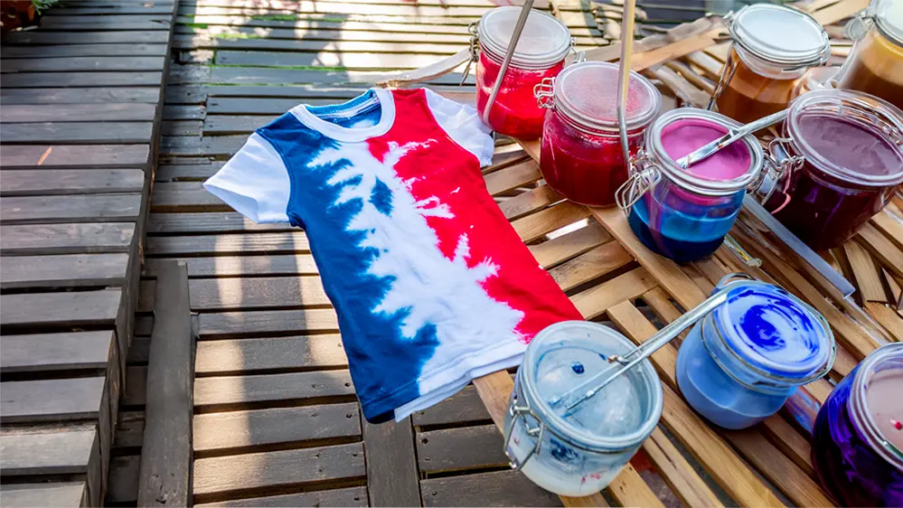 A white shirt with red and blue tie-dye lies on a table that holds several cans of dye and painting tools.