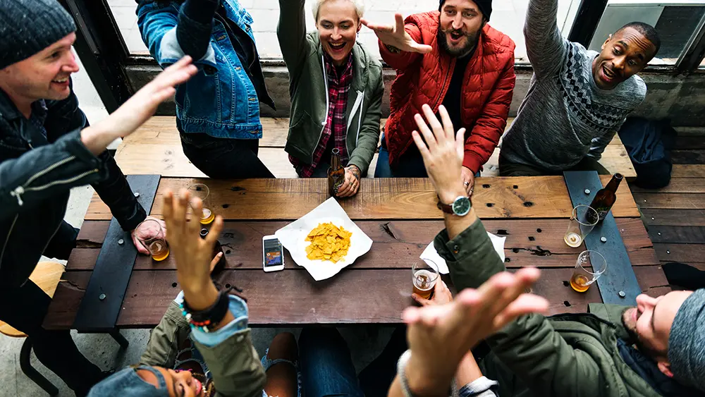Group of people raising their arms jubilantly around a table that holds drinks, chips, and a smartphone. Trivia is an excellent 4th of July game. They’re playing a game of freedom trivia.