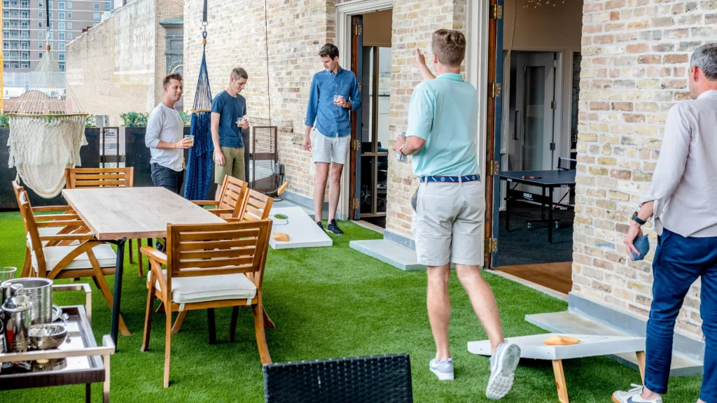 Group of people playing 4th of July games, such as cornhole. They’re playing on a terrace yard which also holds patio furniture. Inside, there’s a ping-pong table.