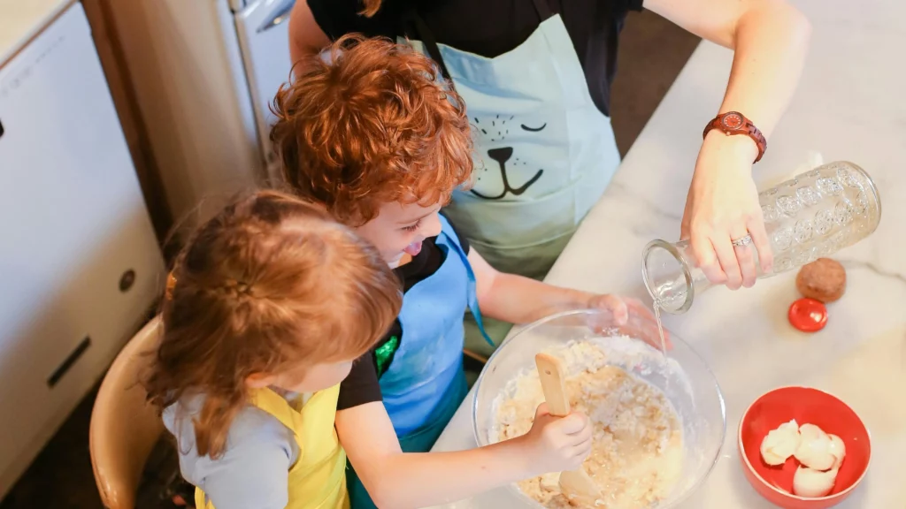 An adult pours a liquid into a bowl containing flour as a young child mixes it and a second child looks at it. They’re baking 4th of July treats together.