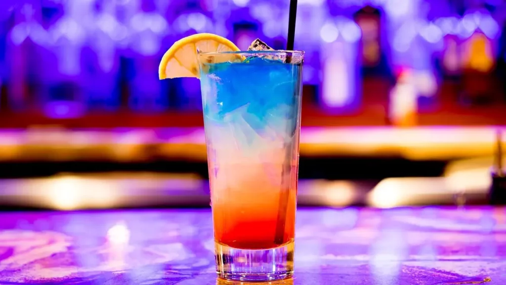 Glass holding a drink with red, white, and blue hues, a black straw and a lemon slice on the edge. It’s a freedom themed cocktail perfect for 4th of July.
