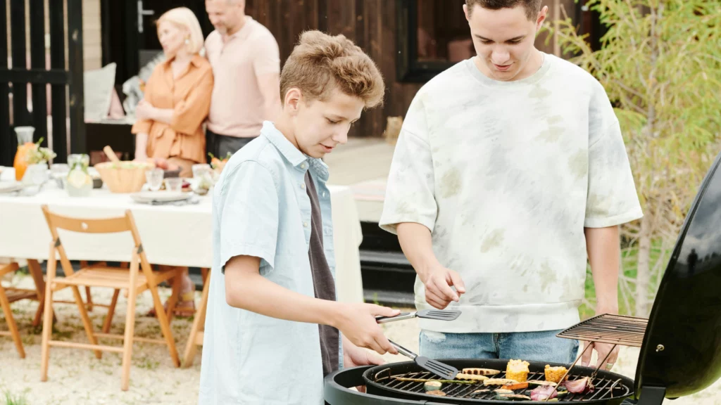 A youth attempting to rearrange food on a grill using tongs while a second person offers direction.