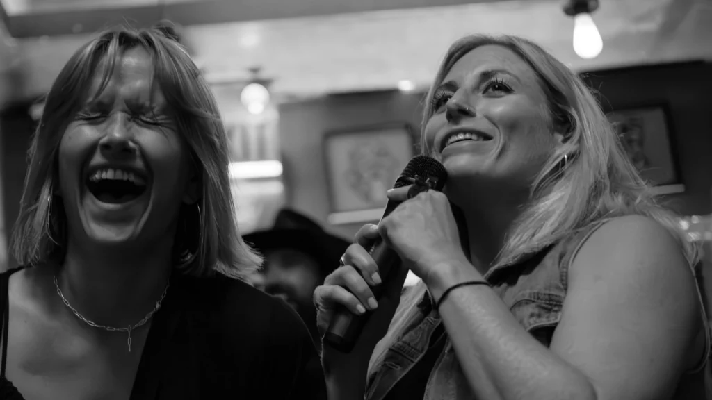 Black and white photo of two women enthusiastically singing patriotic karaoke songs. They are laughing and having a fun time.