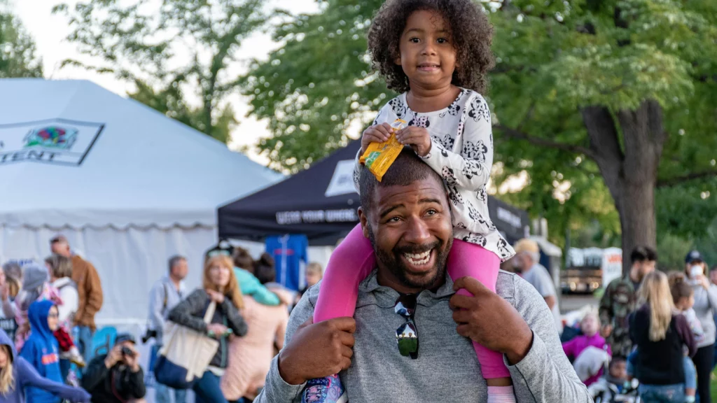 Man carries child on his shoulders against a background of crowd and tents.