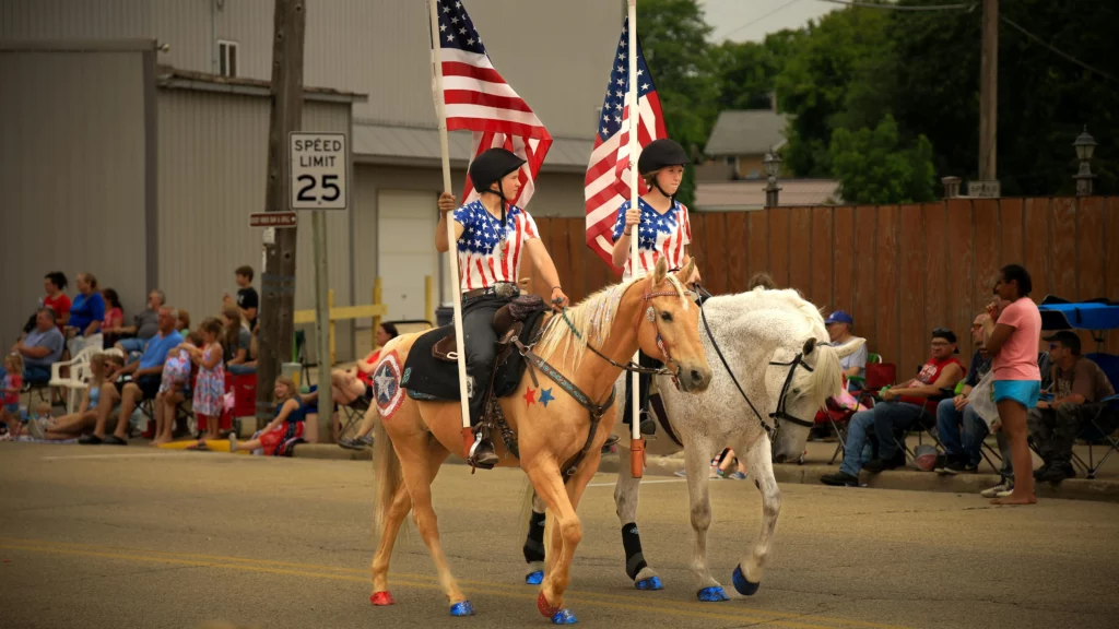 A happy 4th of July parade. Two horses with red, white, and blue accents on their hooves and coat are being rode on a street by two people wearing flag-print shirts while carrying an American flag each. There are spectators at the side of the road.