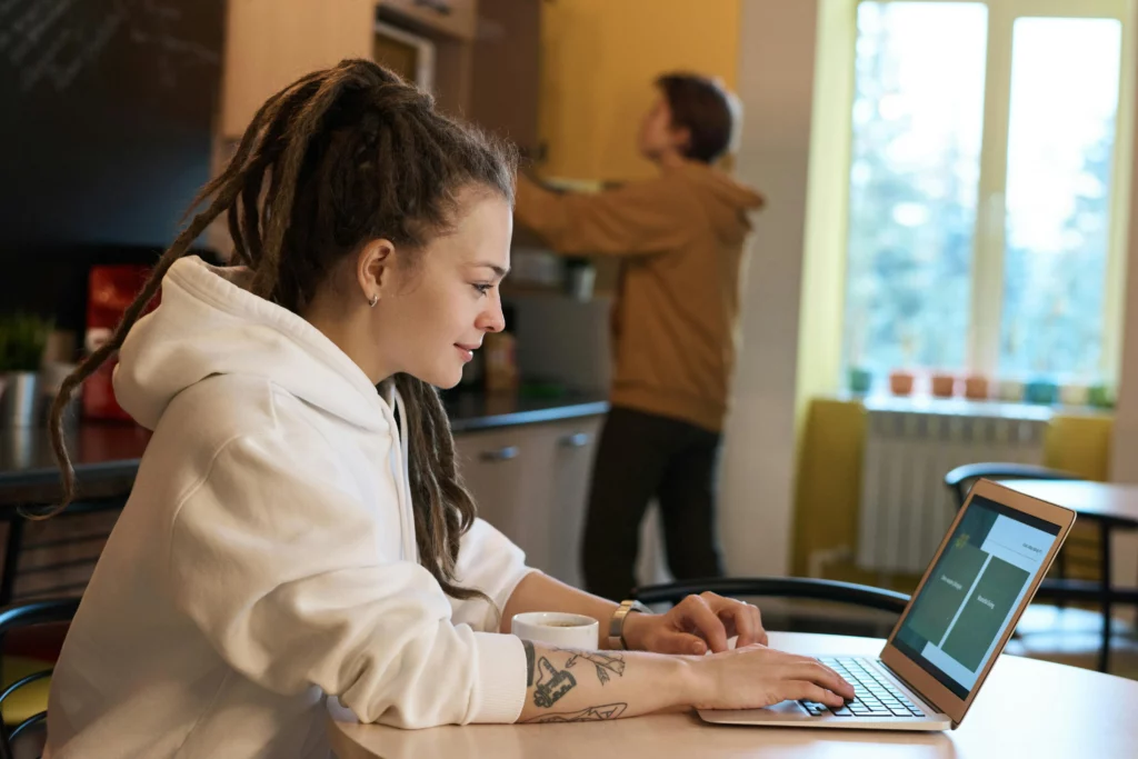 Woman wearing a white hoodie sits at a table and is working a freelance side hustle from a laptop. Meanwhile, a man in seen in the background trying to retrieve something from a cabinet.