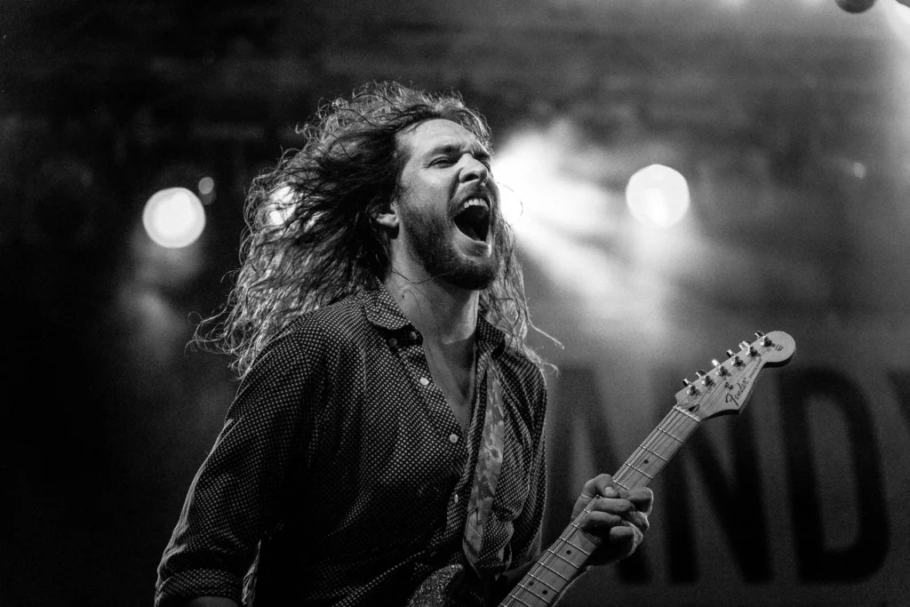 Black and white image of a man with long hair singing while playing a guitar. He works a side hustle as a performing artist. 