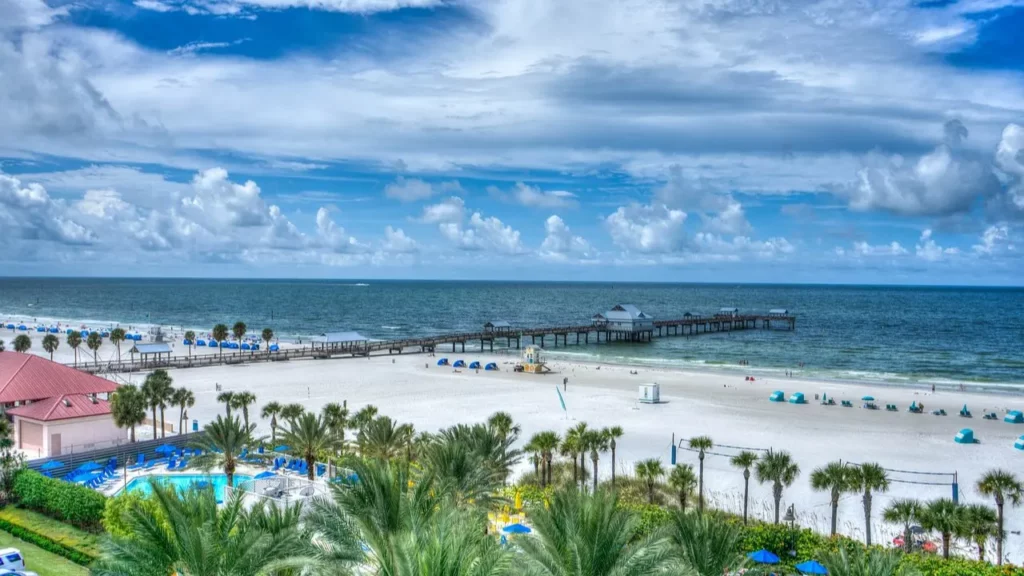 Snapshot of Clearwater Beach in Florida shows the blue sky with several different cloud formations, choppy waters, and a pristine beach dotted with beach umbrellas and chairs. A pool surrounded by lounge chairs and umbrellas can be seen flanked by palm trees all around.