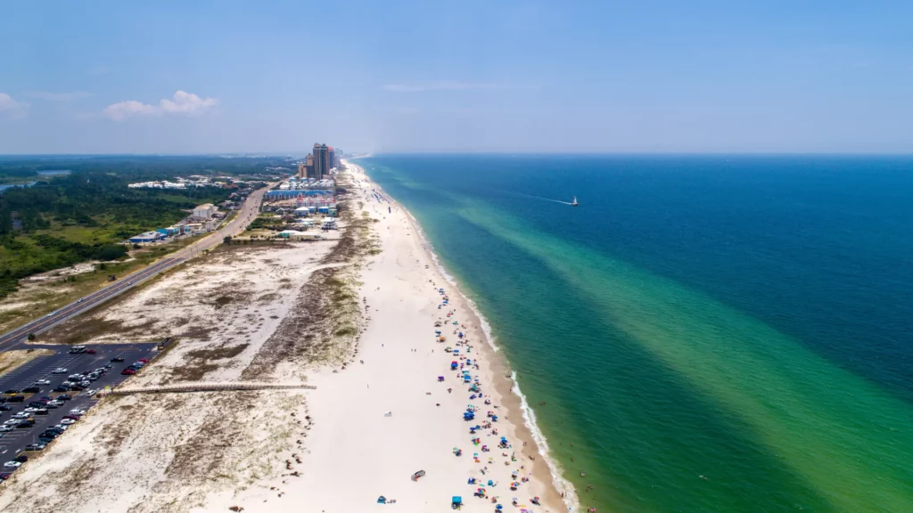 Snapshot of the Alabama Gulf Shores beach along blue-green waters while numerous people are scattered across the sand. There is a parking lot with cars, a few high-rise buildings, and greenery around the beach and a sailboat sails across the waters epitomizing cheap beach vacations. 