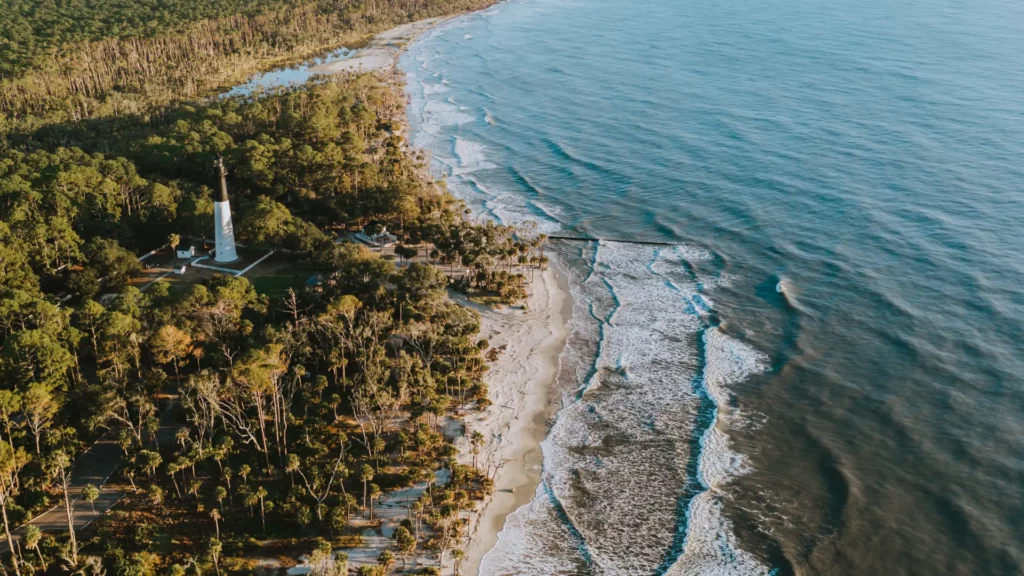 Snapshot of Hunting Island State Park in South Carolina. A sandy beach meets the waters along with an abundance of palm trees. The striking black and white Hunting Island Lighthouse completes the image for cheap beach vacations.