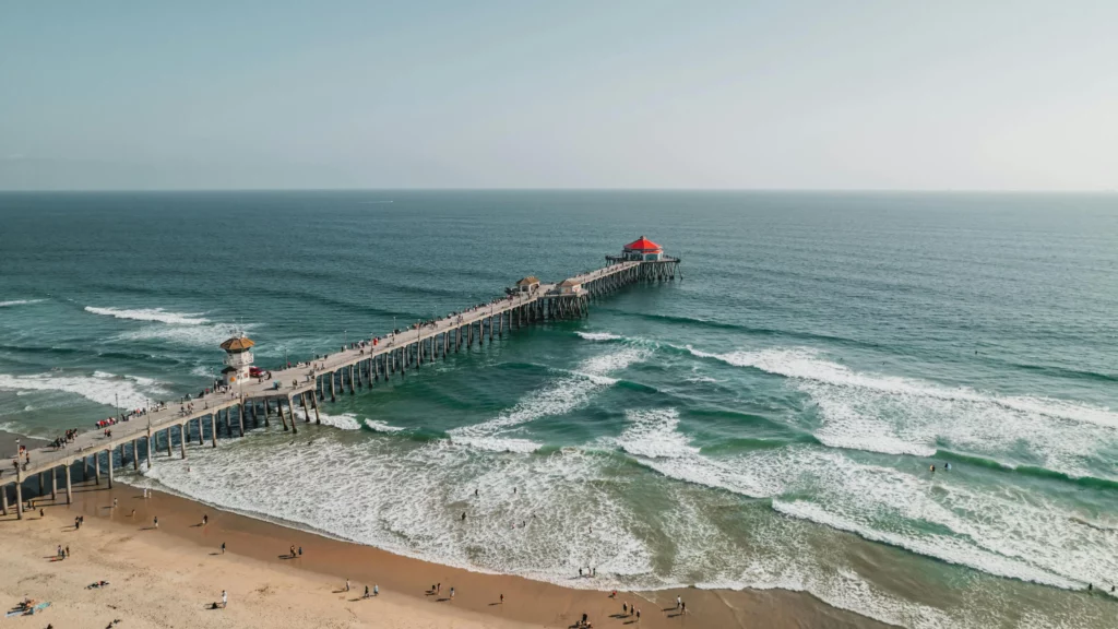Snapshot of Huntington Beach, California. A sandy beach dotted with people against the blue-green waters. A pier juts out from the beach to the water and has people walking on it.