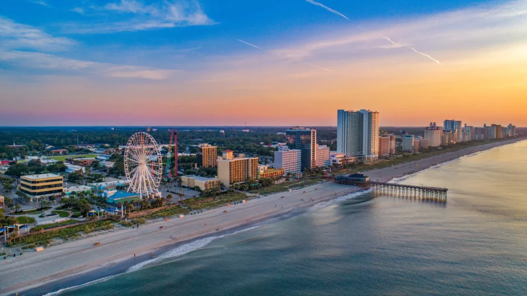 Snapshot of Myrtle Beach in South Carolina. Tranquil waters meet the sandy beach while a pier emerges into the water. A tall white Ferris wheel can be seen on the beach in addition to several high-rise buildings.