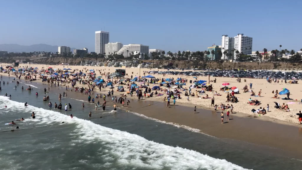 Snapshot of California’s Pacific Beach scattered with people on the sand and in water, possibly enjoying affordable beach vacations. There are palm trees and buildings along the horizon.