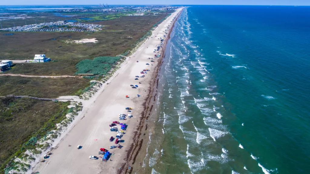 Snapshot of the Texas Port Aransas beach and waters along with multiple cars in the sand as people are enjoying cheap beach vacations. Buildings and other structures can also be seen.
