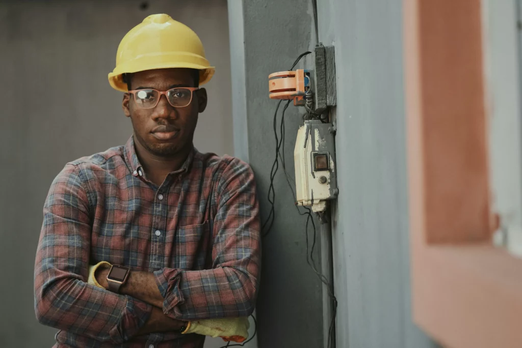 An electrician wearing a yellow helmet, glasses, safety gloves, a watch, and a plaid shirt is looking at the camera while leaning against a wall and keeping his arms folded across his chest. Electrical equipment and wires are seen protruding from the wall