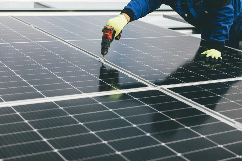 Image of a solar panel installer wearing blue outerwear and hands wearing florescent yellow and black safety gloves. The person is working with an electric tool on solar panels.