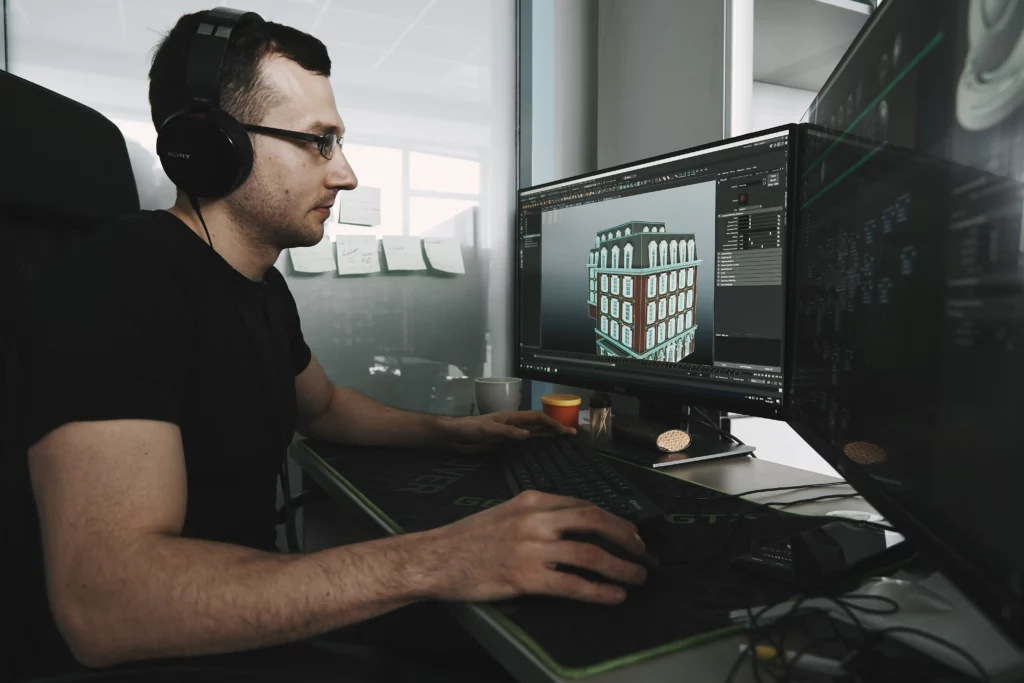 Image of a web developer. He is wearing a black short-sleeved t-shirt and glasses working at a desktop with two monitors.