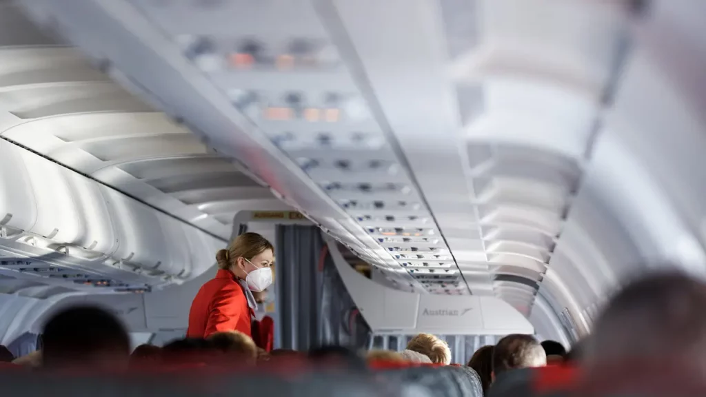 View of an aircraft cabin with passengers seated while a flight attendant wearing a red coat and face mask is seen standing.